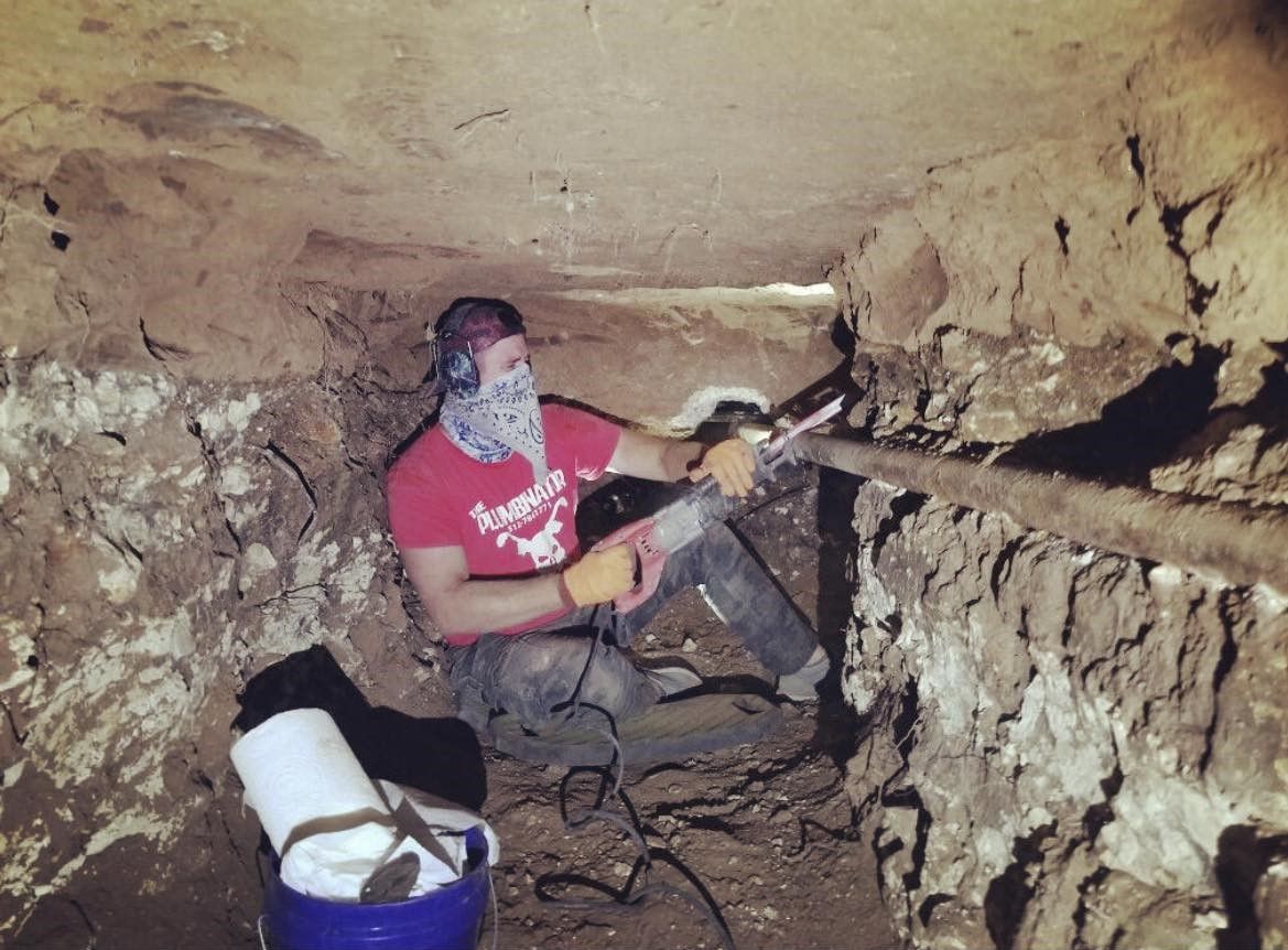 Worker in a narrow tunnel drilling rock, with tools and a bucket nearby.