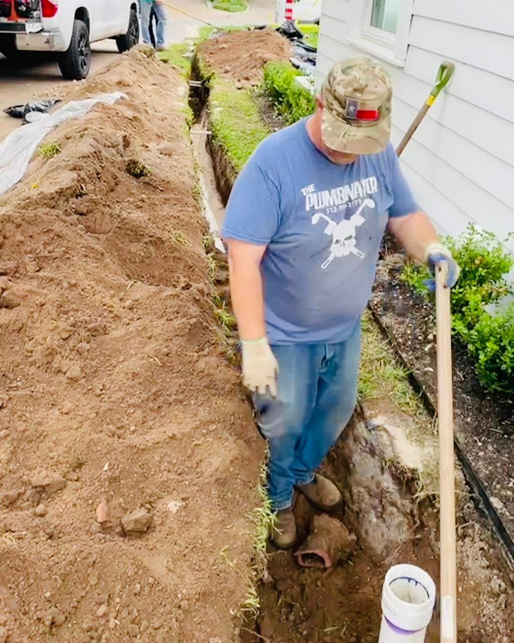 Worker in blue shirt digging a trench beside a house with a shovel and pipe.