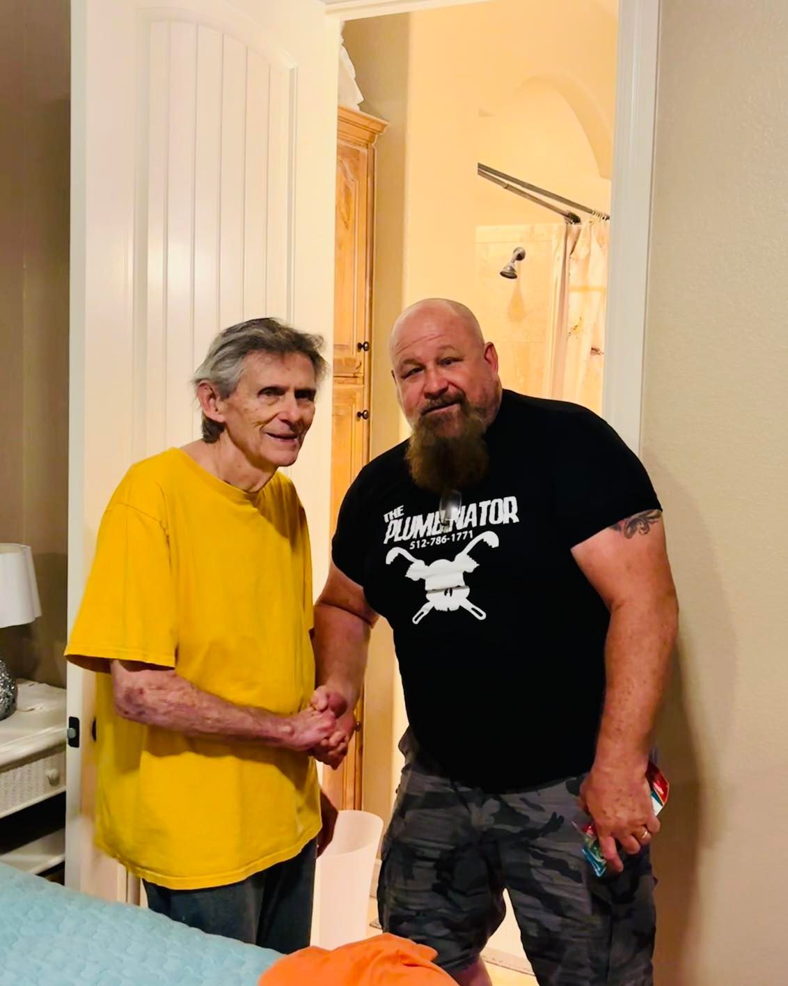 Two men standing together indoors, one in a yellow shirt and the other in a black T-shirt, smiling at the camera.