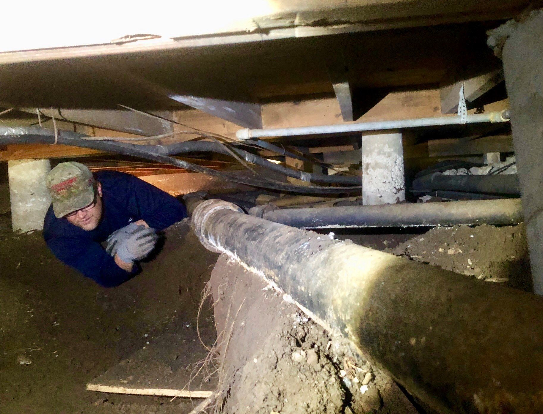Worker crawling under a low wooden structure in a dusty crawlspace, inspecting beams and pipes.