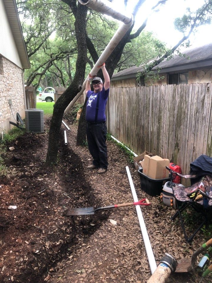 Person holding a large tree limb in a narrow backyard beside a fence, with yard tools on the ground.