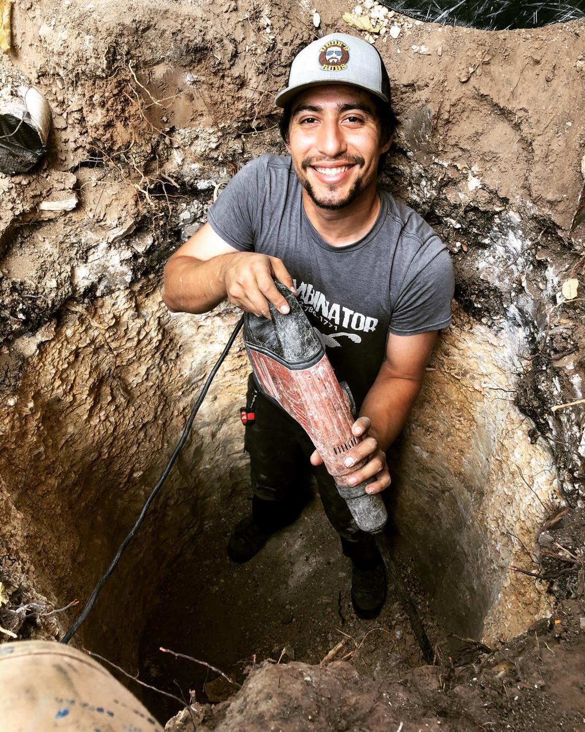 Man in a hard hat smiling while holding a large pipe inside a muddy excavation pit