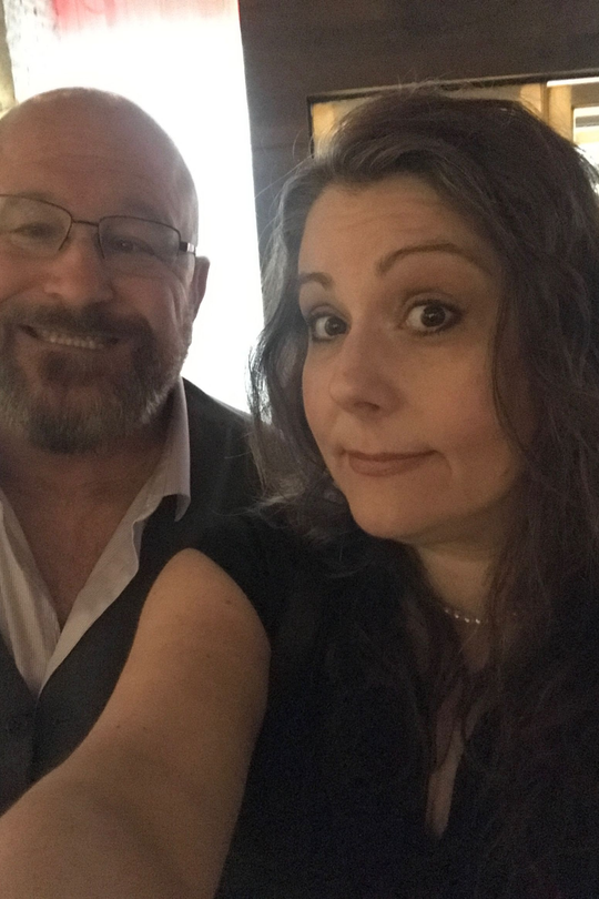 Two people posing for a selfie indoors, one smiling and one with a raised eyebrow.