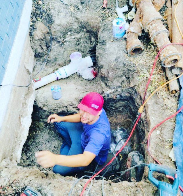 Worker in a red cap emerging from a deep trench beside exposed pipes and muddy ground