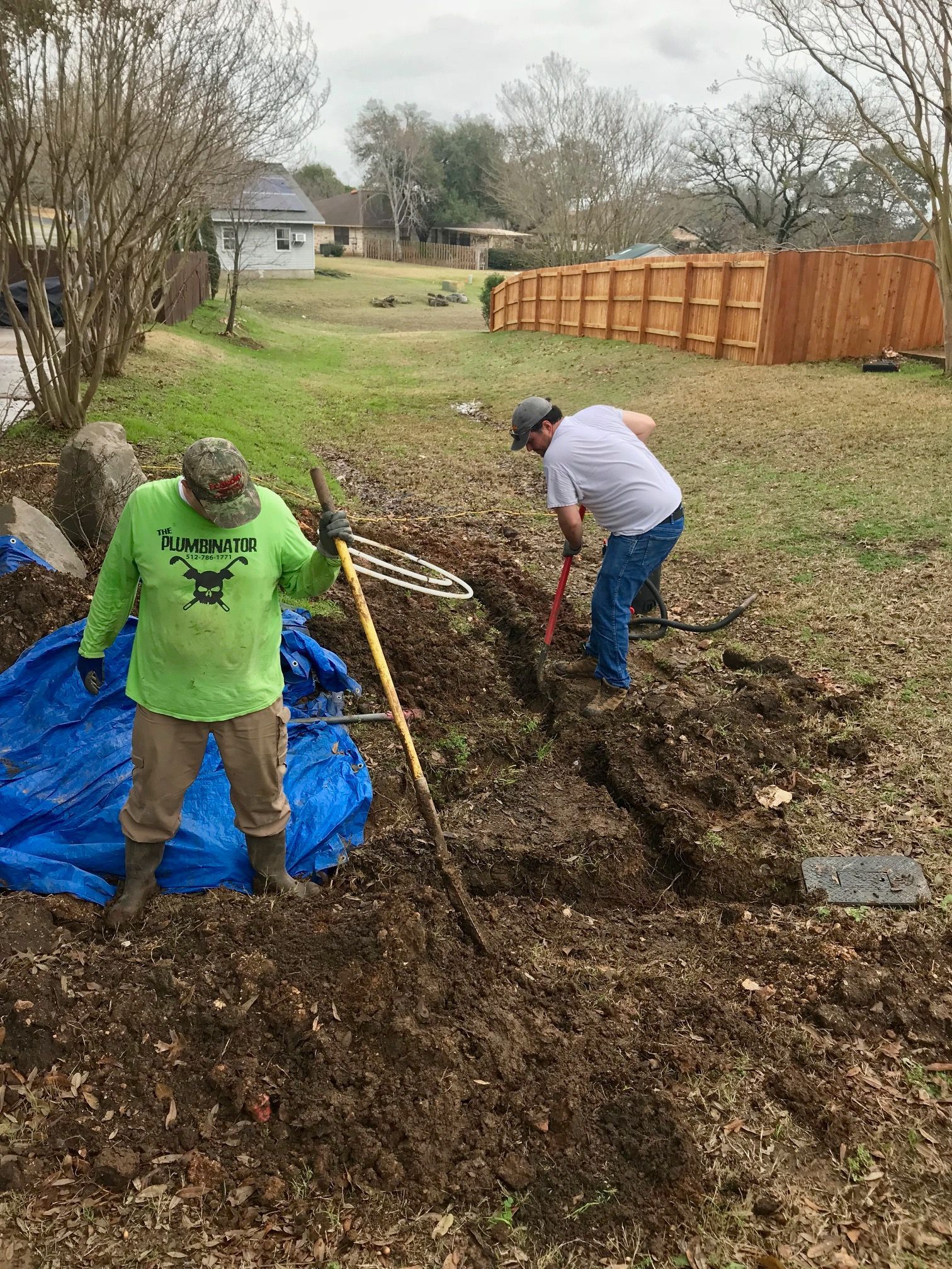 Two people raking leaves in a yard beside a blue tarp and wooden fence