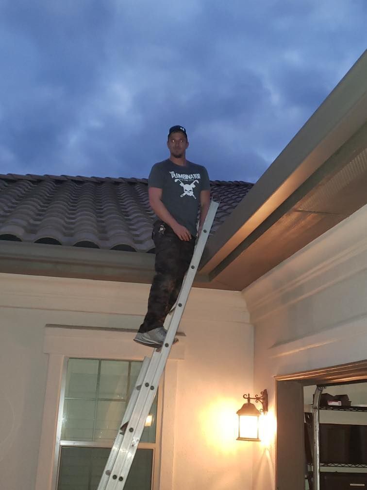 Person standing on a ladder beside a house roof at dusk, holding a tool near the eaves.