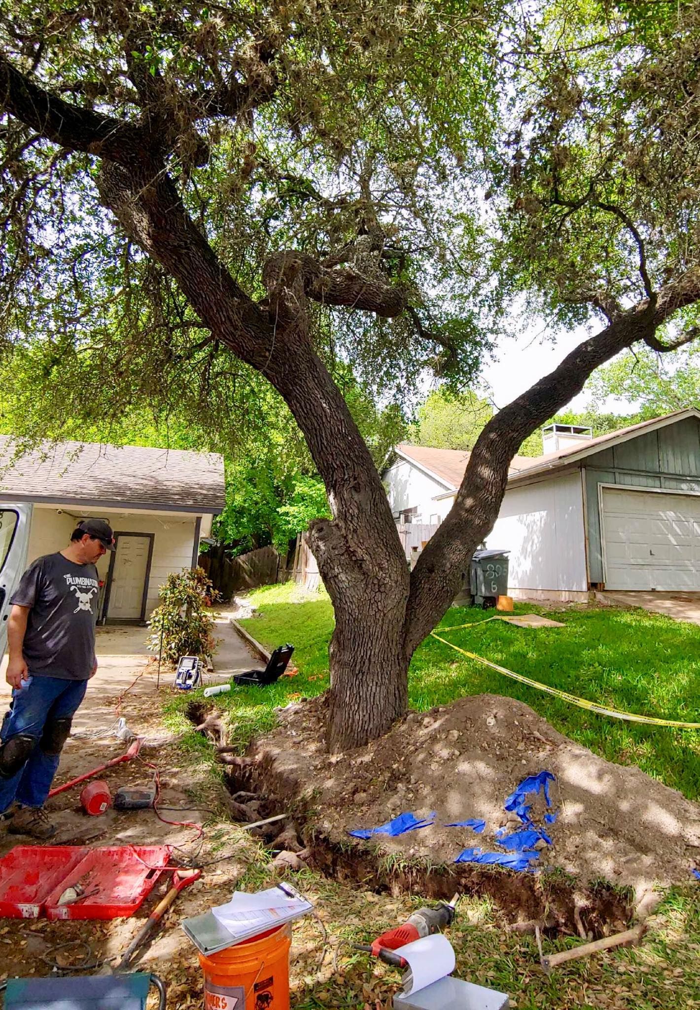 Backyard with a large tree, scattered debris, and a person standing near a garage under cloudy daylight