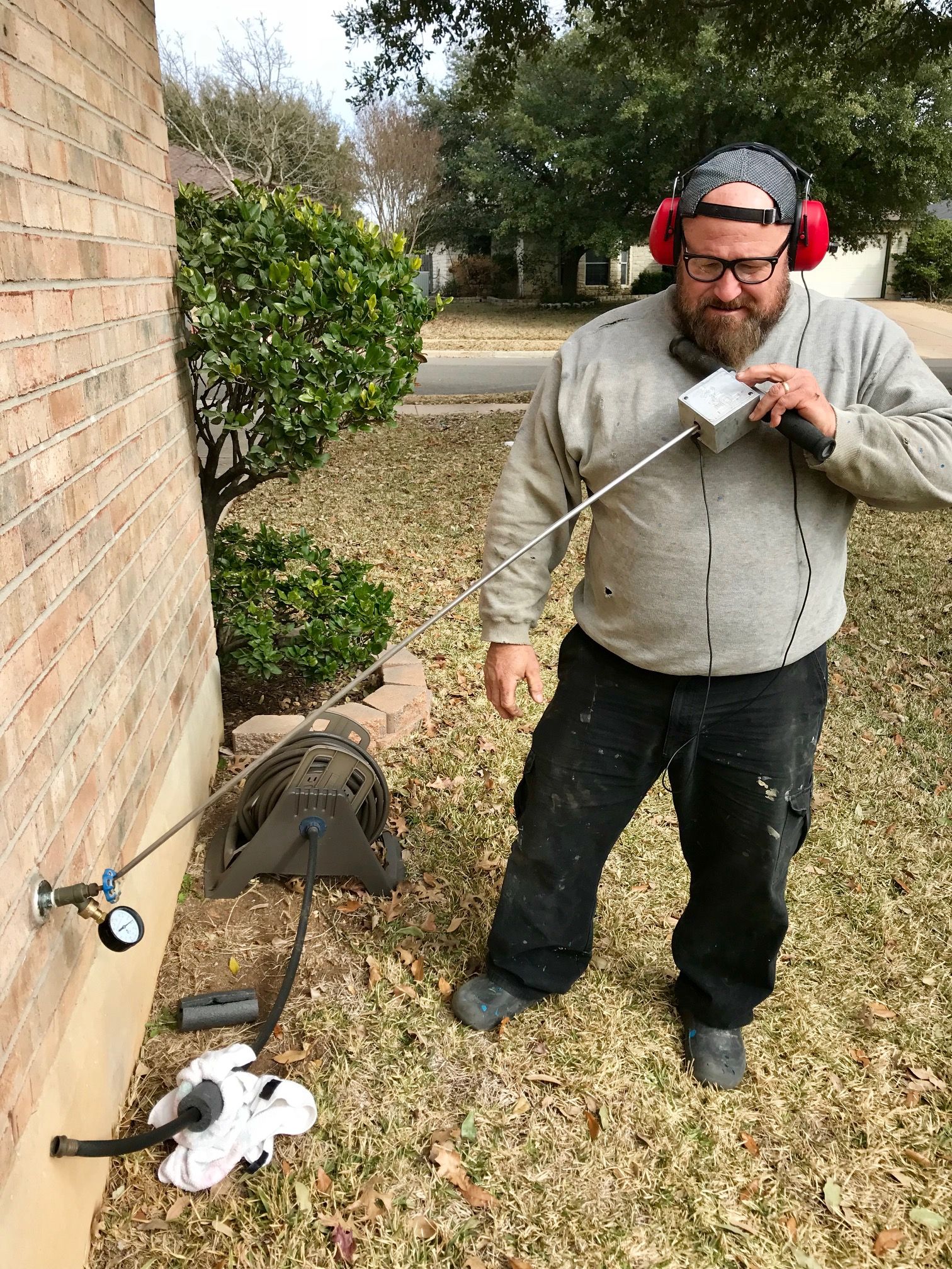 Man using a metal detector near a brick wall, with headphones and a digging tool beside him outdoors.