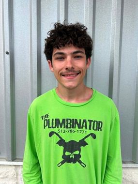 Smiling person in a neon green “The Pluminator” shirt standing before a corrugated metal wall