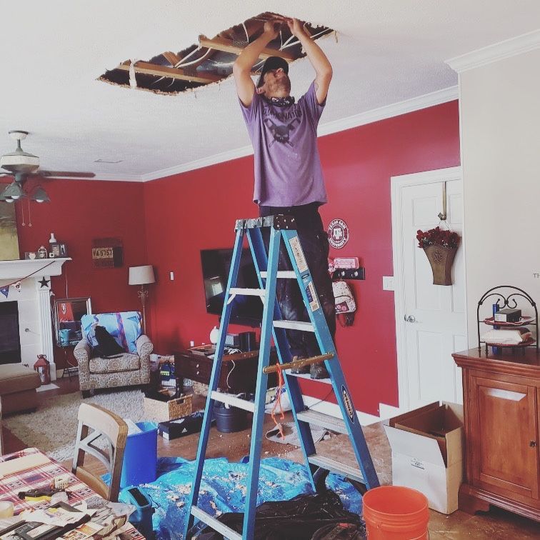 Person on a ladder repairing a damaged ceiling in a red living room.