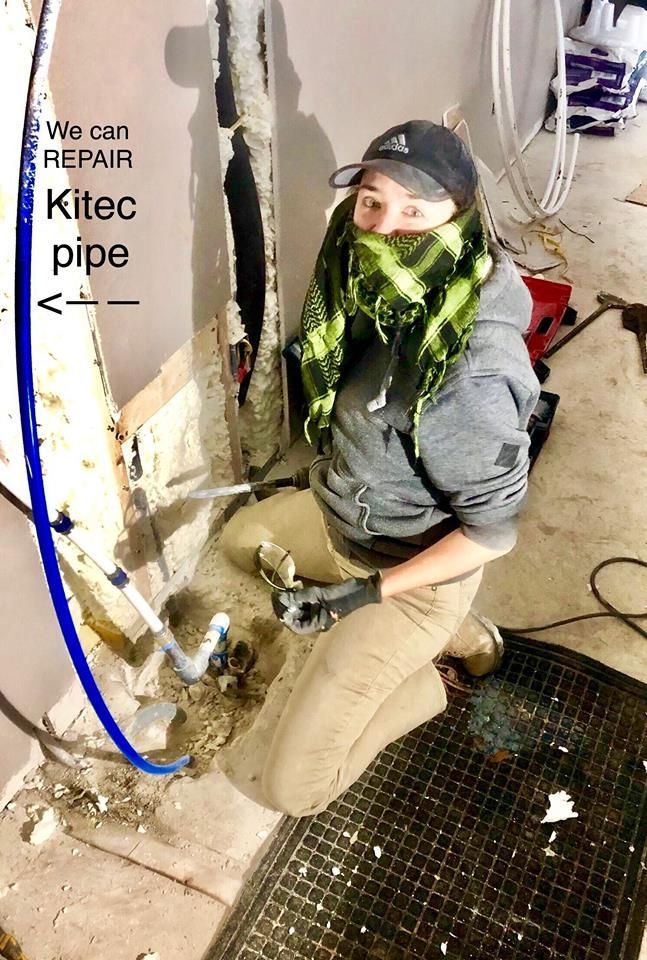 Worker kneeling by a wall repair pipe in a utility room, with green debris on their jacket.