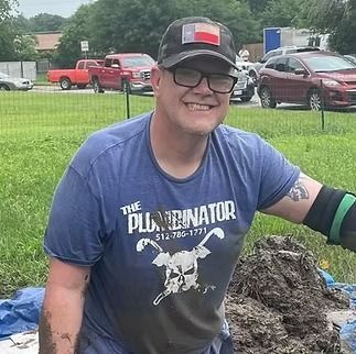 Smiling person in a blue shirt and black cap standing outdoors on grass near vehicles and a rock pile