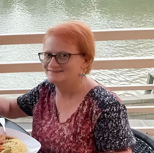 Smiling person with glasses seated at a lakeside table, wearing a red patterned top.