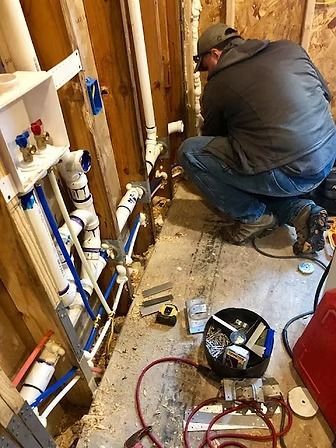 Plumber kneels beside exposed pipes and tools in a narrow utility room during repair work