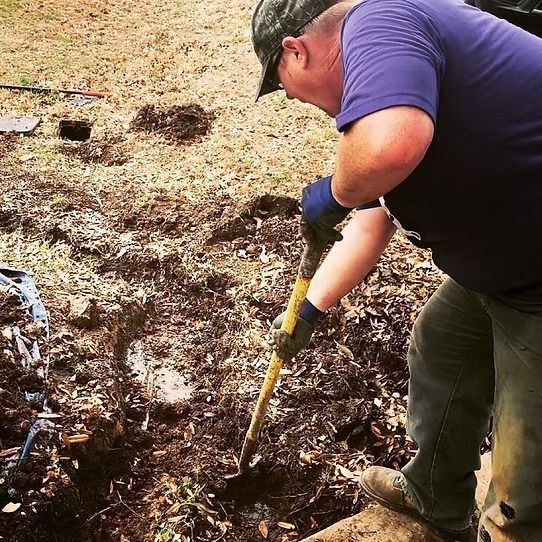 Man digging in a muddy field with a shovel, wearing a cap, purple shirt, and gloves