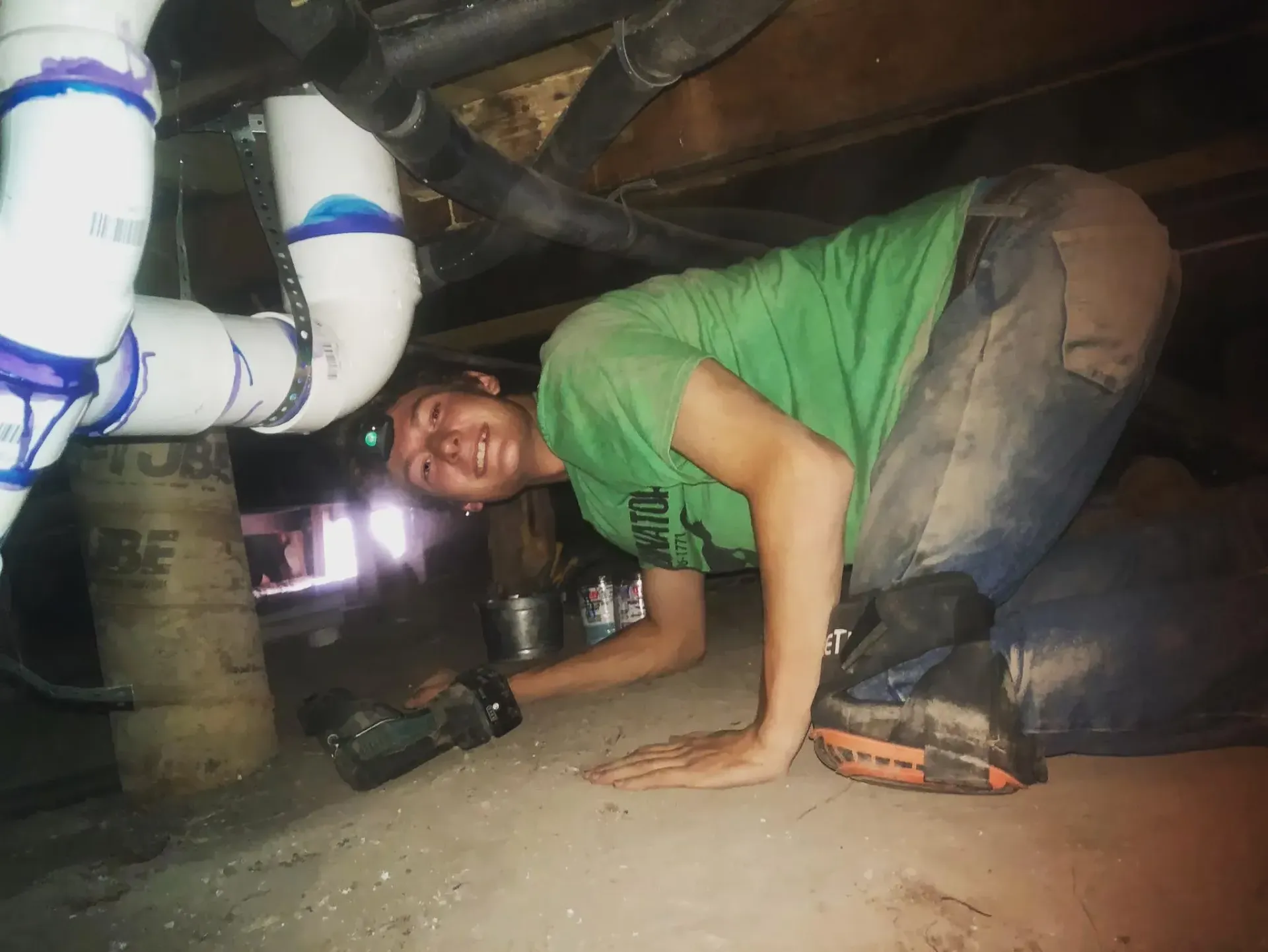 Person crouching in a crawl space beside white pipes, smiling toward the camera.