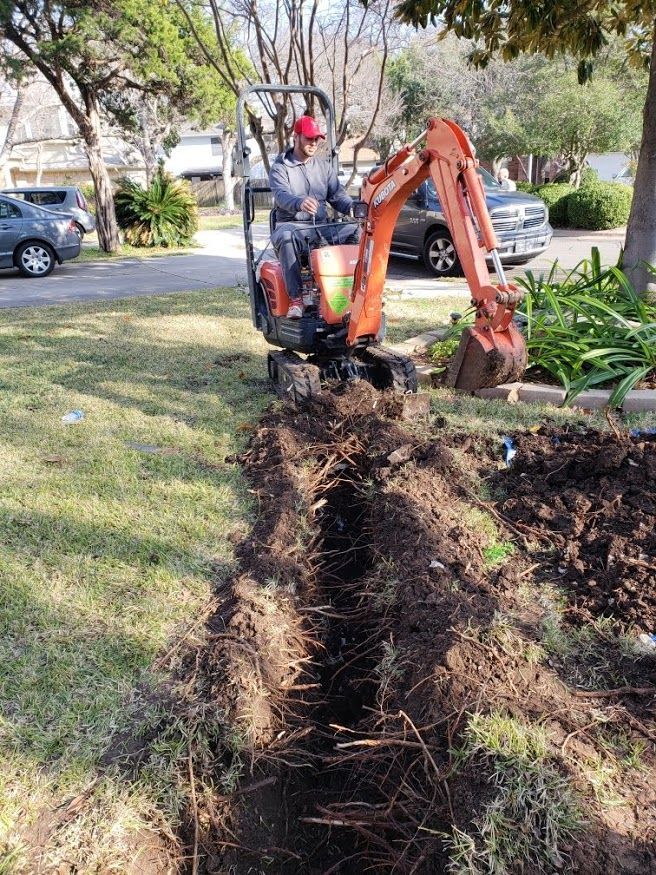 Backhoe excavating a long trench in a grassy yard beside a house and parked cars