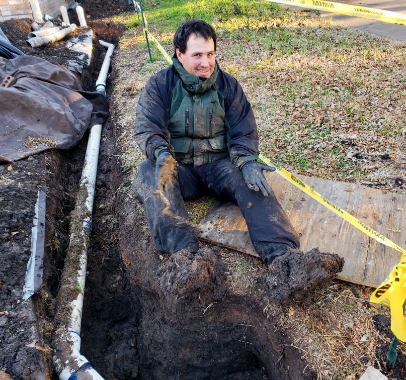 Person sitting in a muddy trench beside yellow caution tape and pipes at an outdoor work site