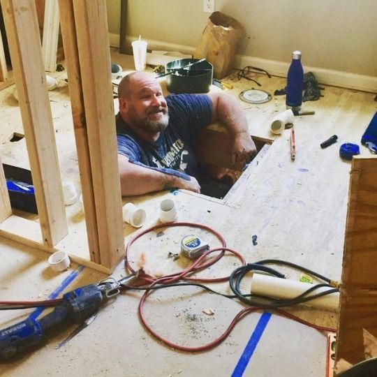 Man working in a partly built room, surrounded by tools and lumber near a floor opening.