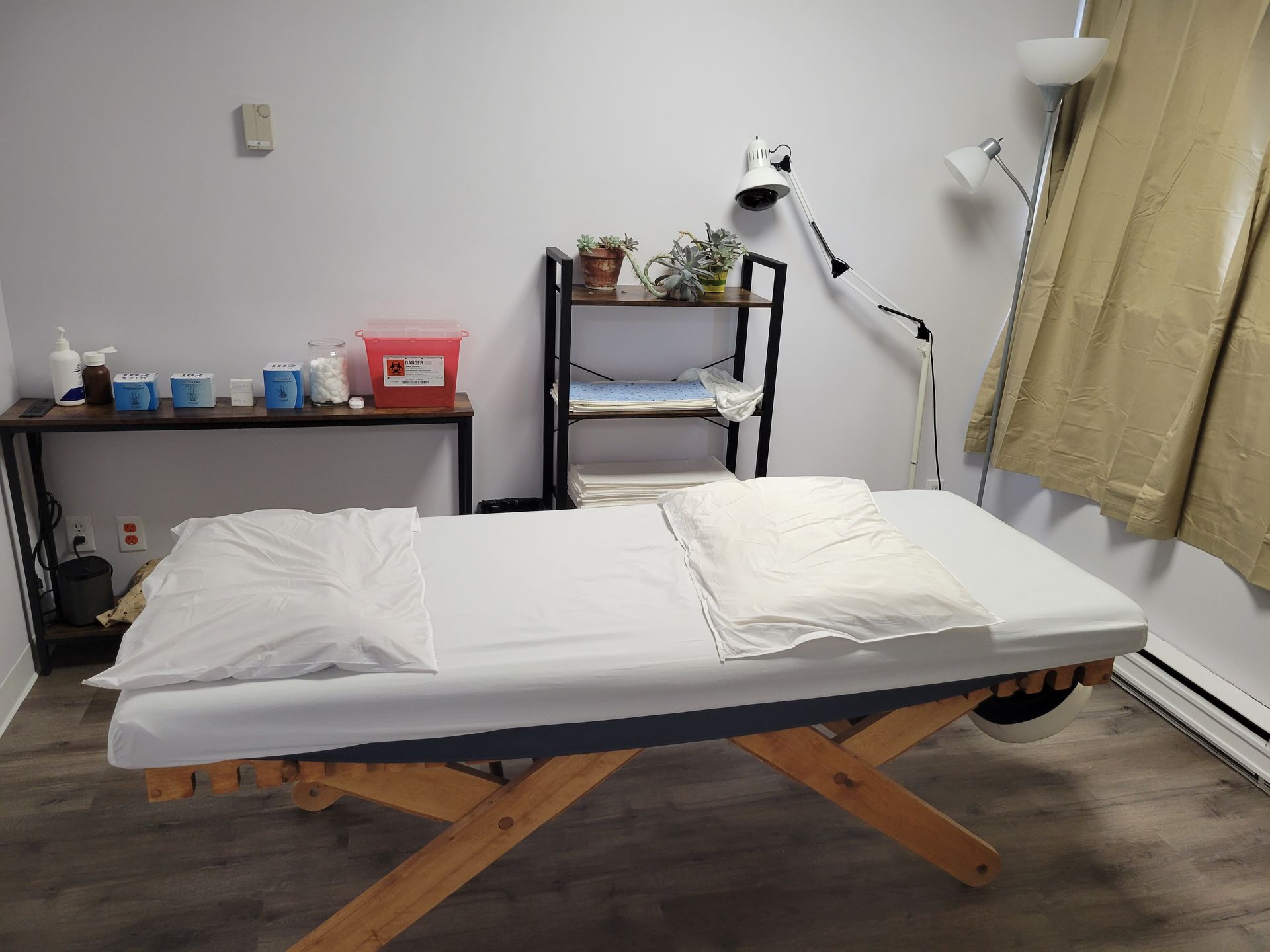Table de massage dans une salle de soins avec serviettes pliées, étagères et lampe près du rideau.