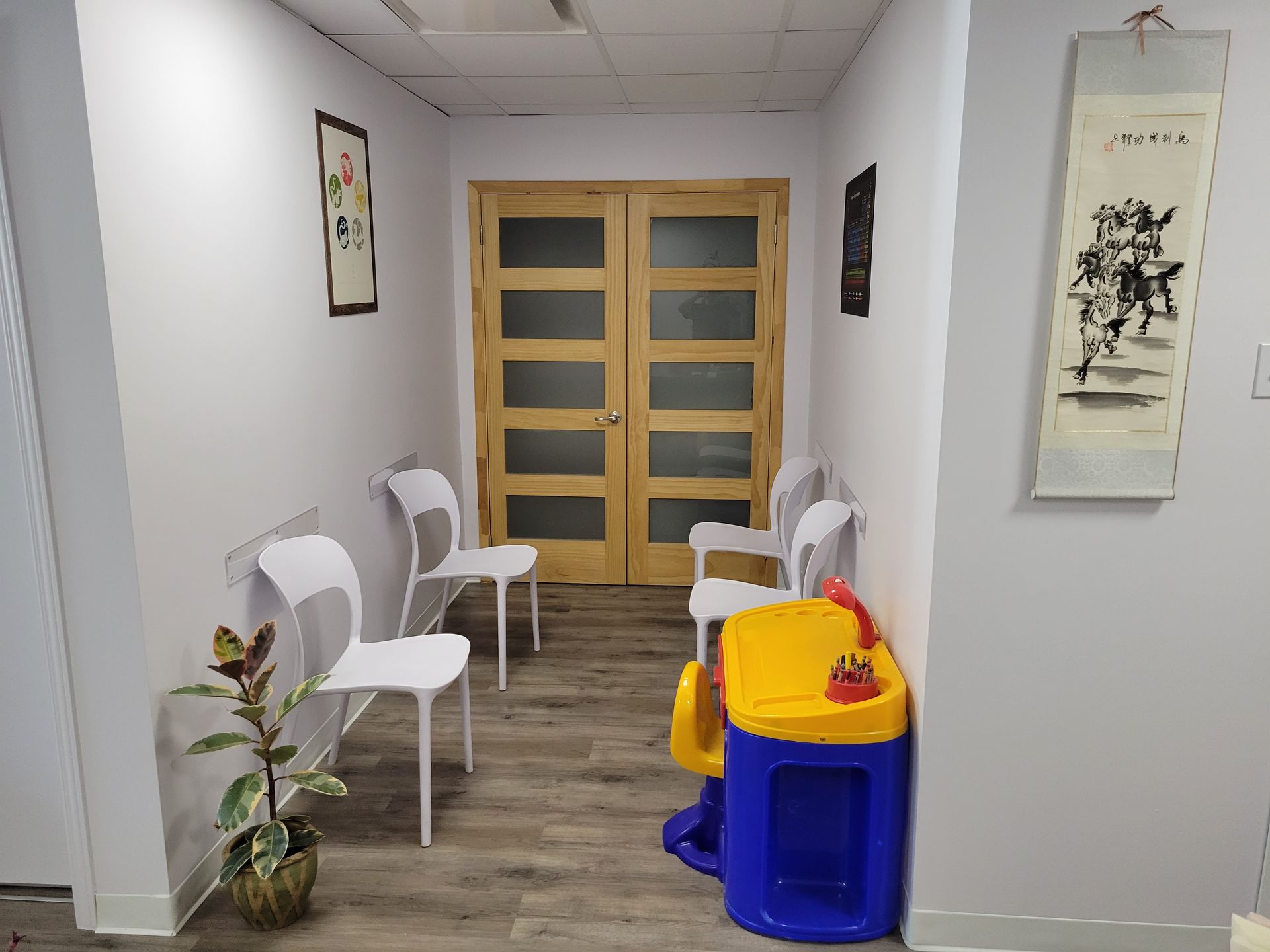 Couloir tranquille avec chaises blanches, plante en pot, décoration murale et petite table à jouets jaune-bleu.