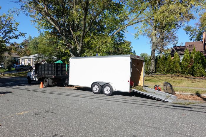 White utility trailer parked on a street beside a black trailer and trees in bright sunlight