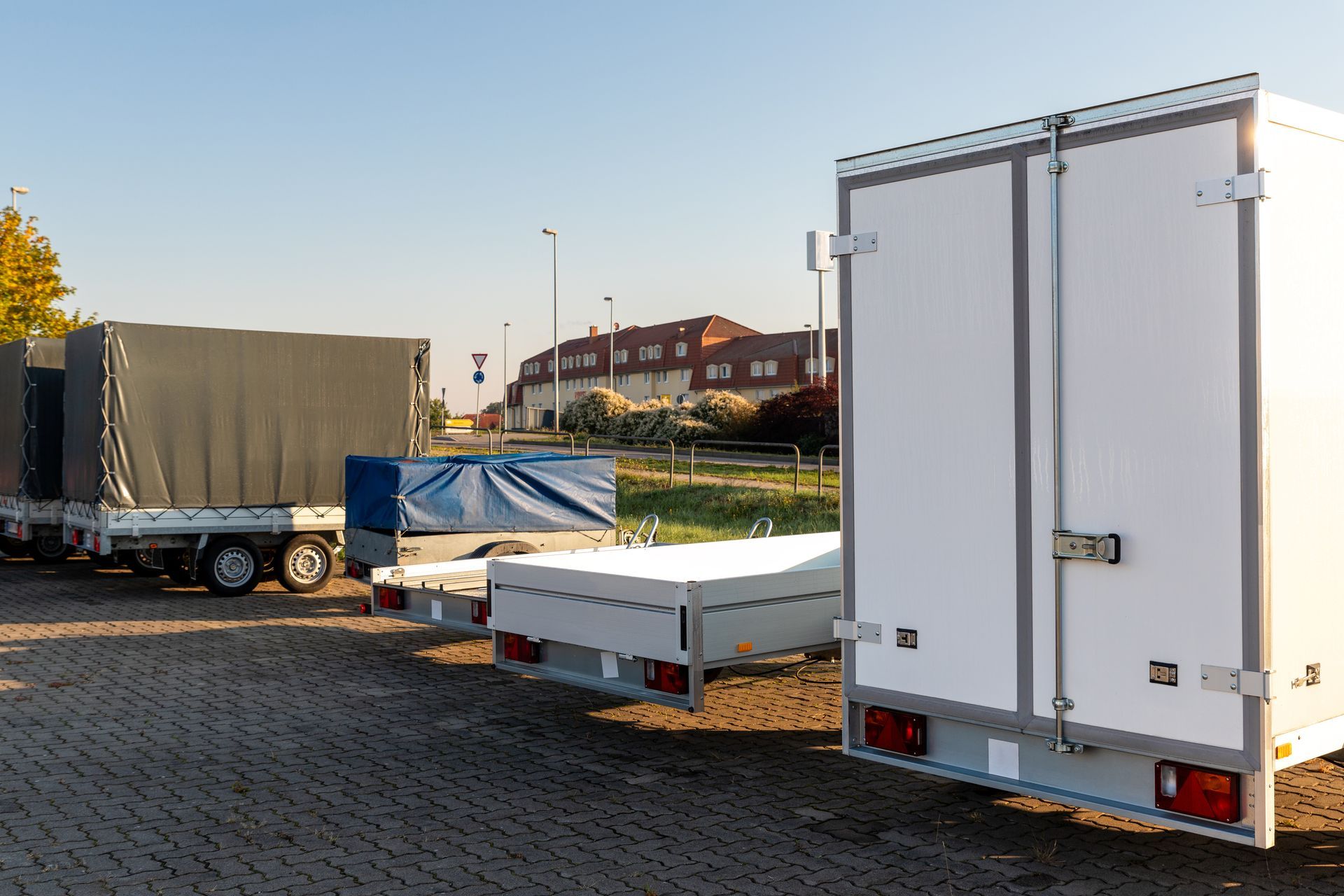 Several empty utility and box trailers parked on a paved lot near a road and residential buildings. Several empty utility and box trailers parked on a paved lot near a road and residential buildings.