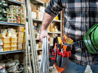Electrician holding wires, wearing tool belt and plaid shirt, in a hardware store.