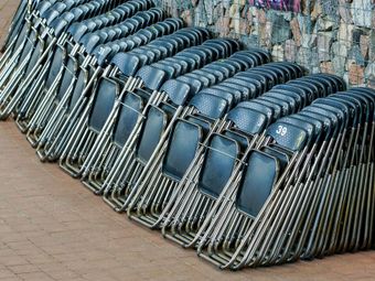 Rows of stacked black folding chairs against a textured brick wall and ground.