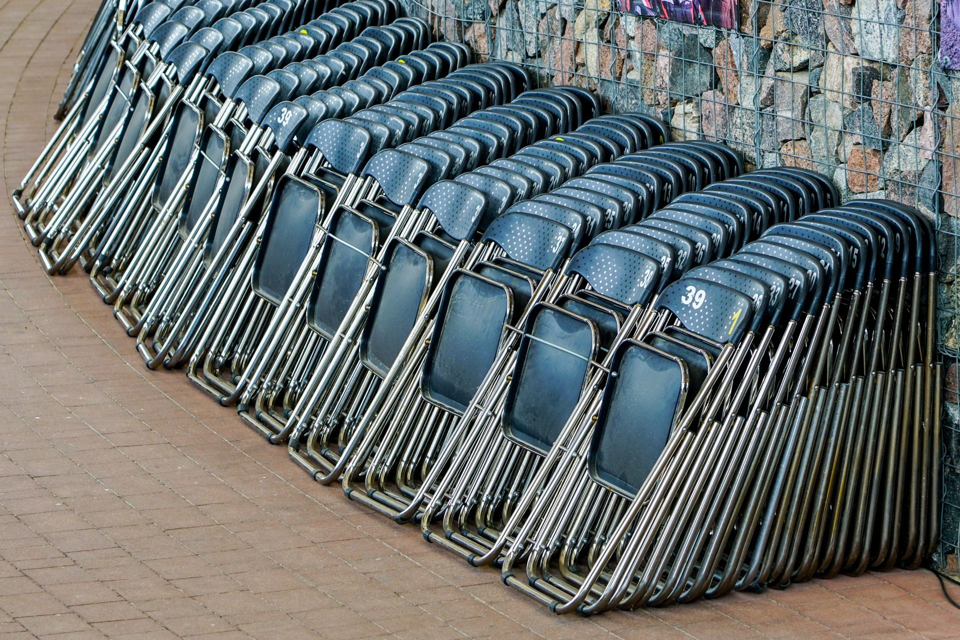 Rows of black folding chairs stacked against a textured stone wall on a sidewalk.