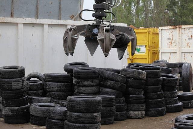 Un artiglio meccanico solleva pneumatici da un grande mucchio in una discarica, in una giornata nuvolosa.