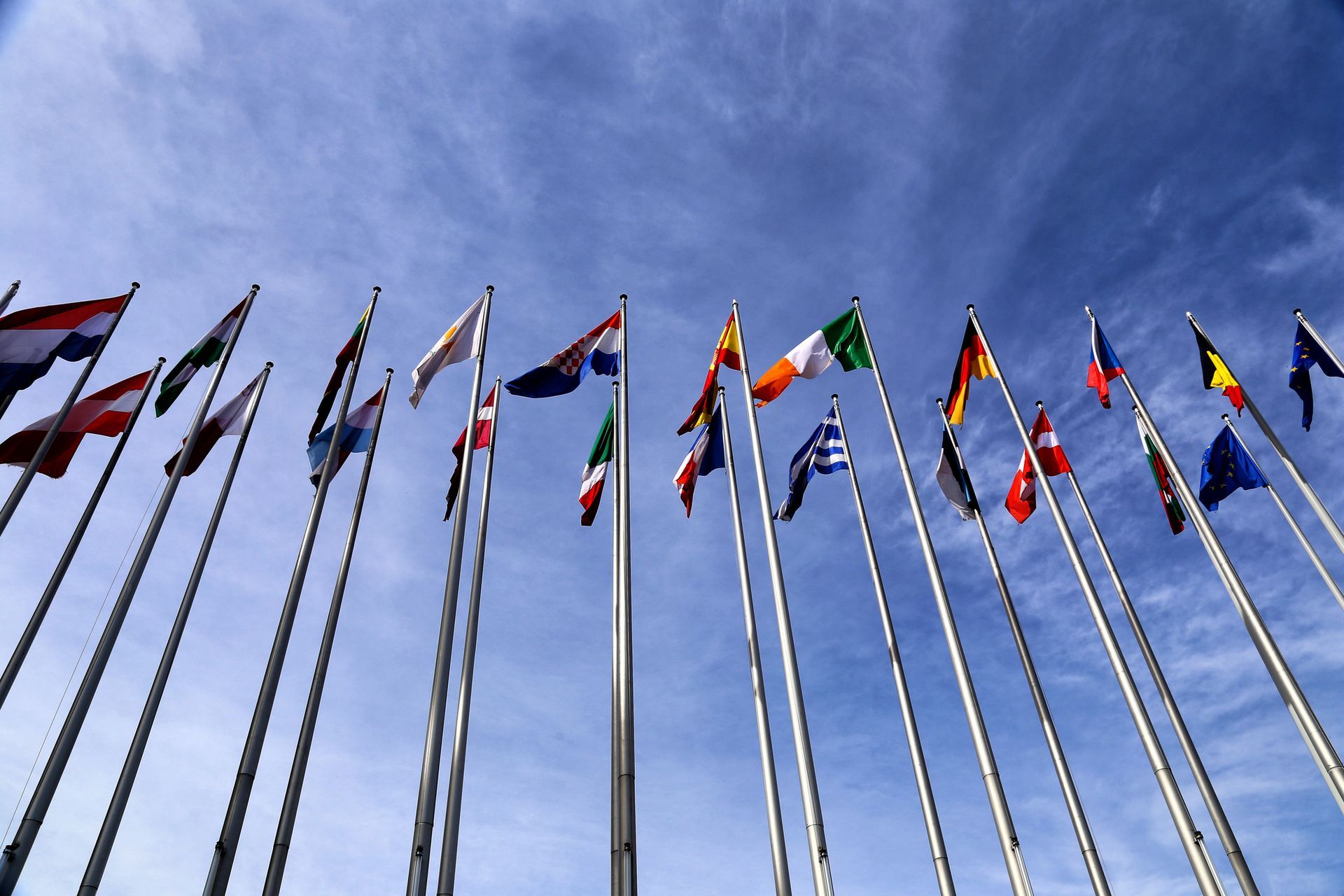 A low-angle shot of many colorful national flags flying on tall poles against a blue sky with light clouds.