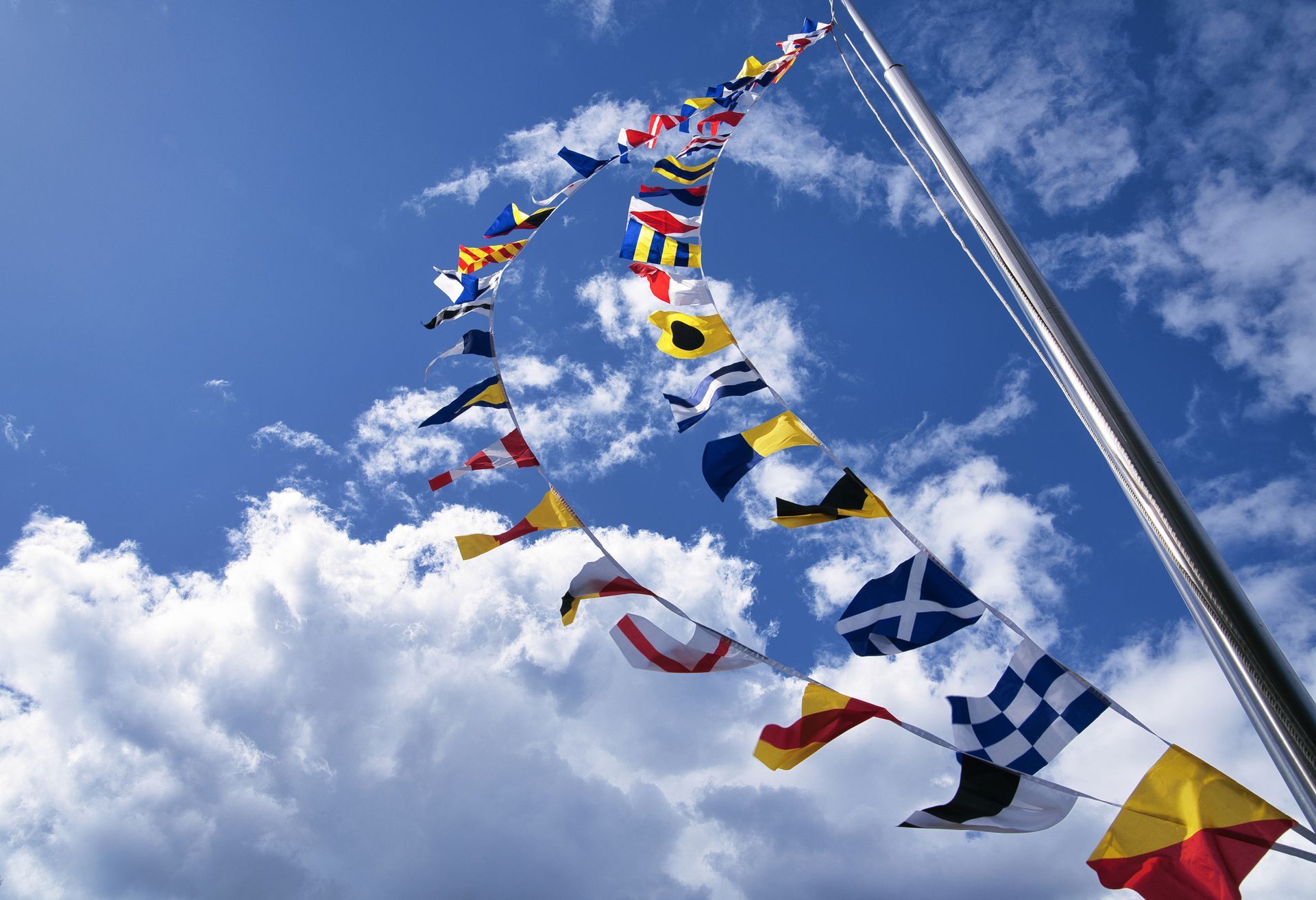 A long string of colorful nautical signal flags flying from a mast against a blue, cloudy sky.