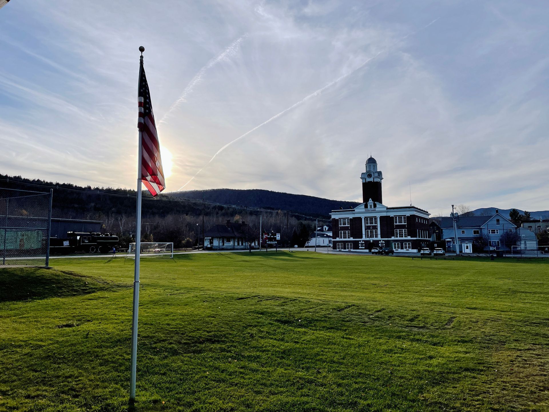 An American flag flies over a grassy field toward a town hall building under a sunny, late afternoon sky.