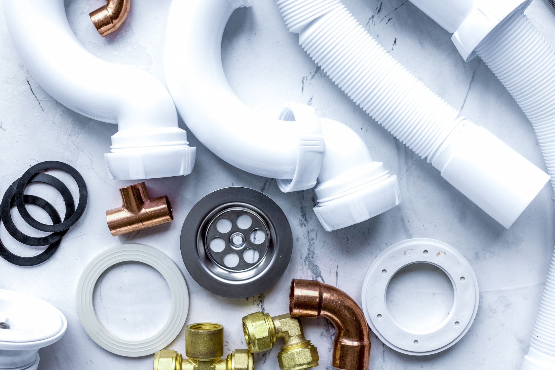 White and copper plumbing parts, including pipes, fittings, and drain parts, arranged on a light surface.