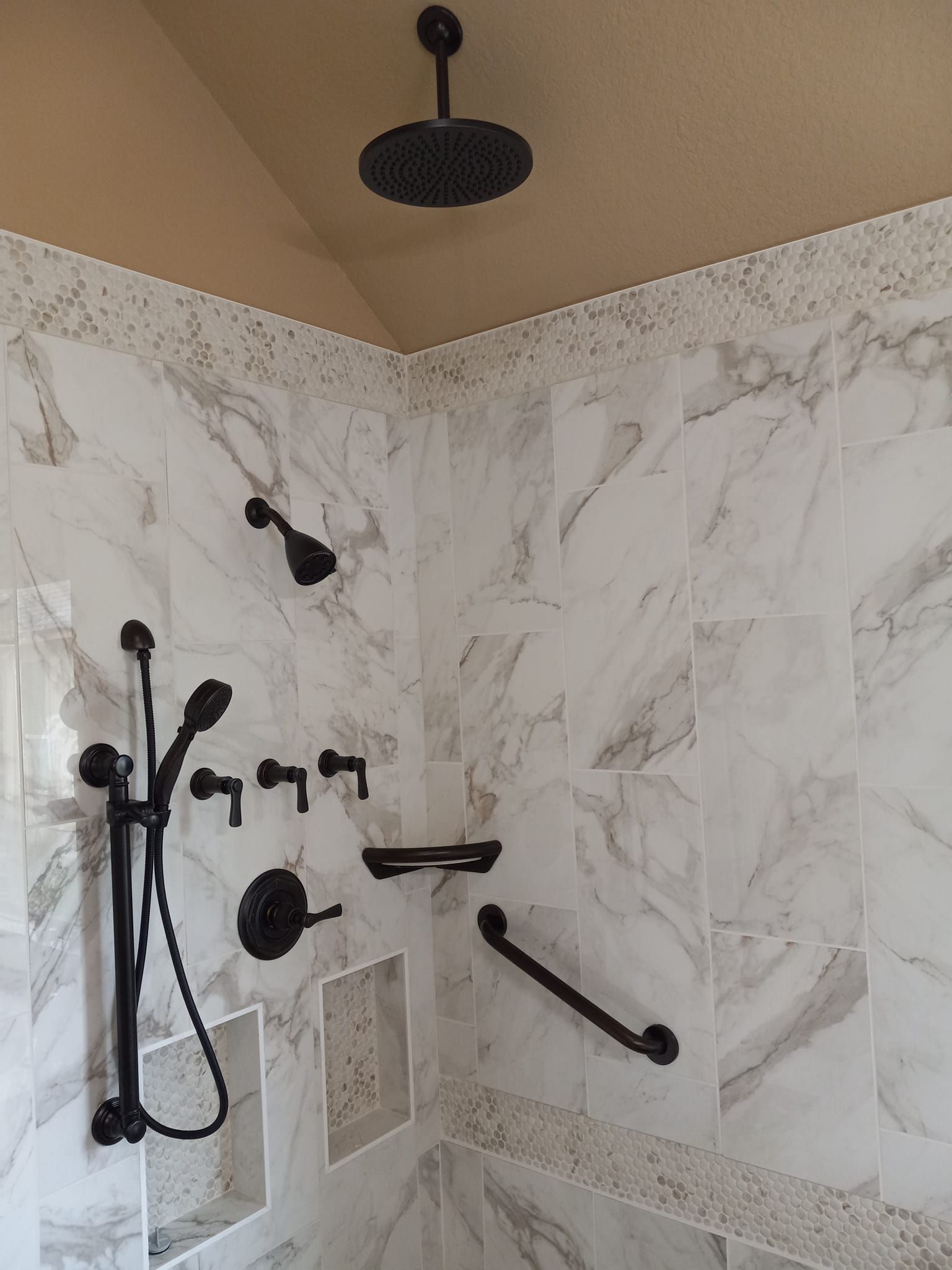 Black shower fixtures in a tiled shower with a grab bar, rain shower head, and recessed shelf.