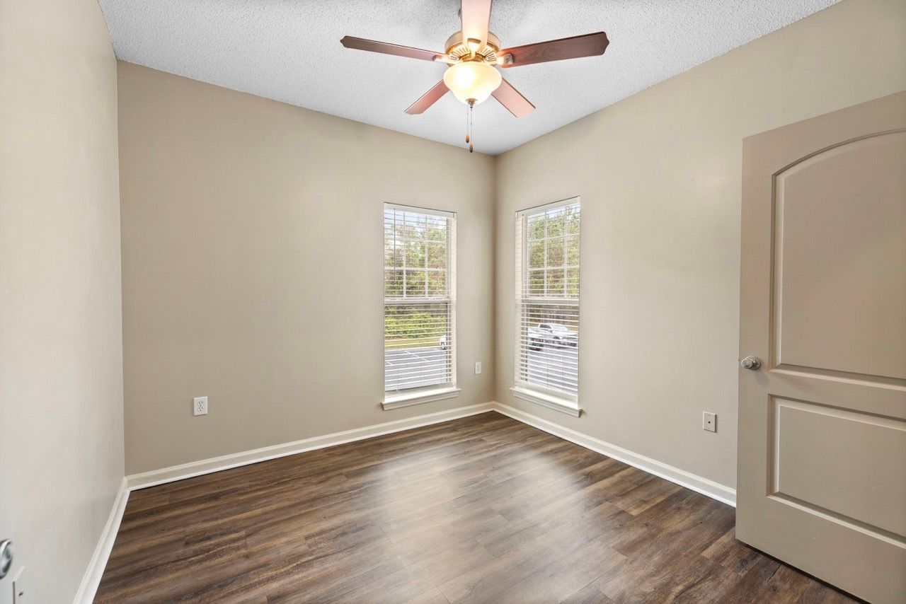 Empty apartment room with beige walls, two windows with blinds, wood-look flooring, and a ceiling fan.