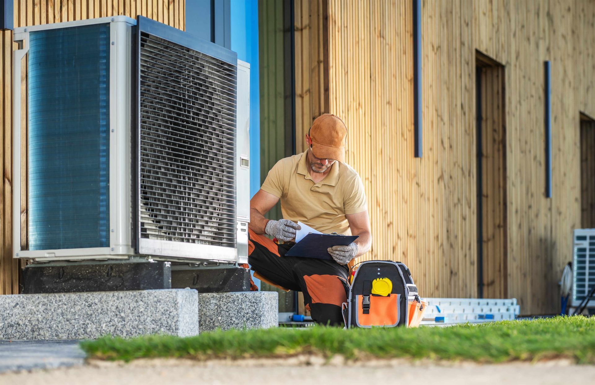 HVAC technician inspecting an air source heat pump outside a wood-sided building.