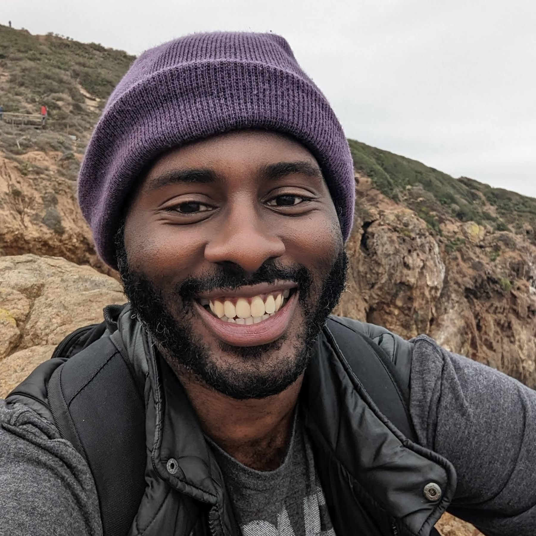 Man with a purple beanie smiles at the camera on a rocky hillside; gray jacket, green and tan cliffs in the background.