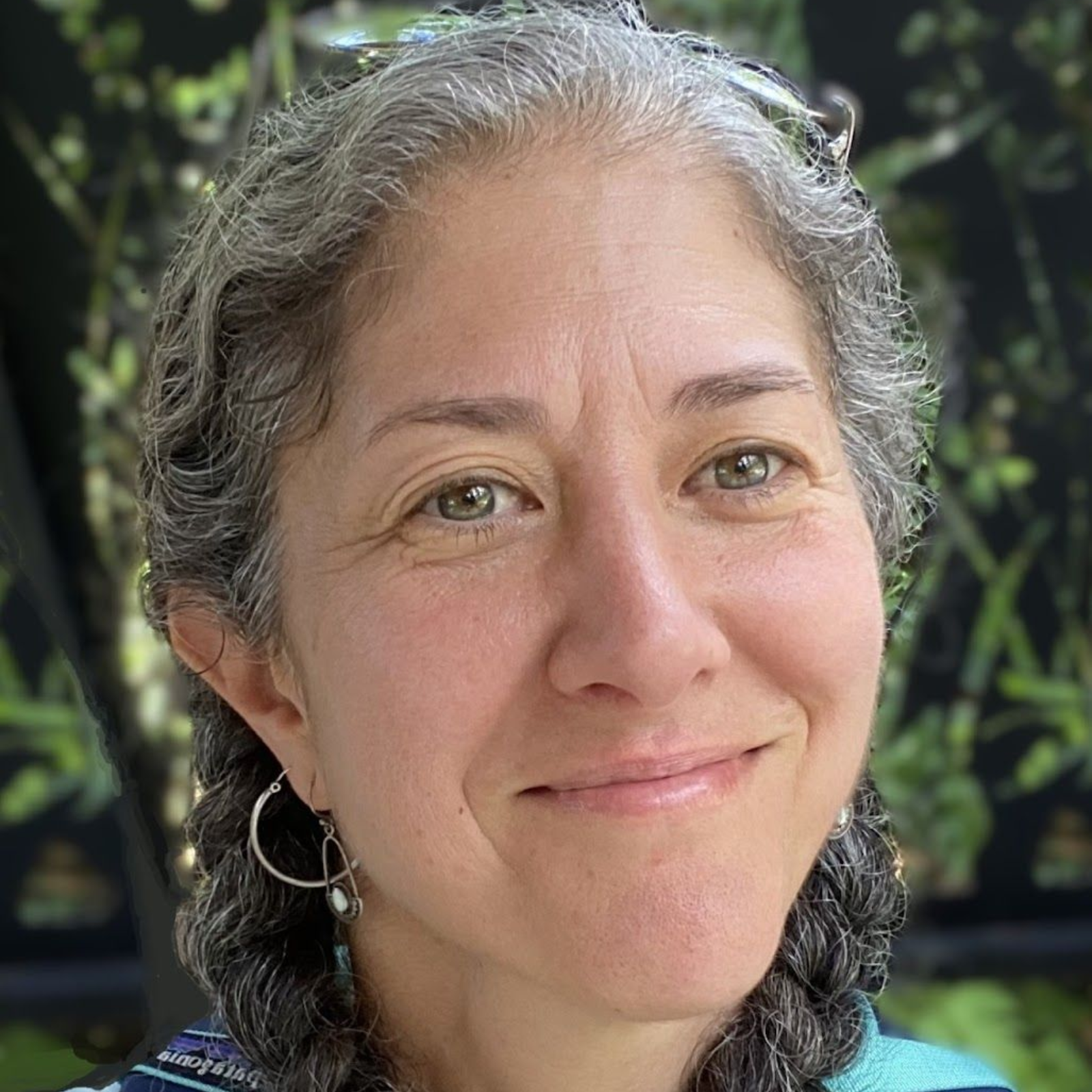 Woman with graying hair smiles outdoors, wearing silver hoop earrings.