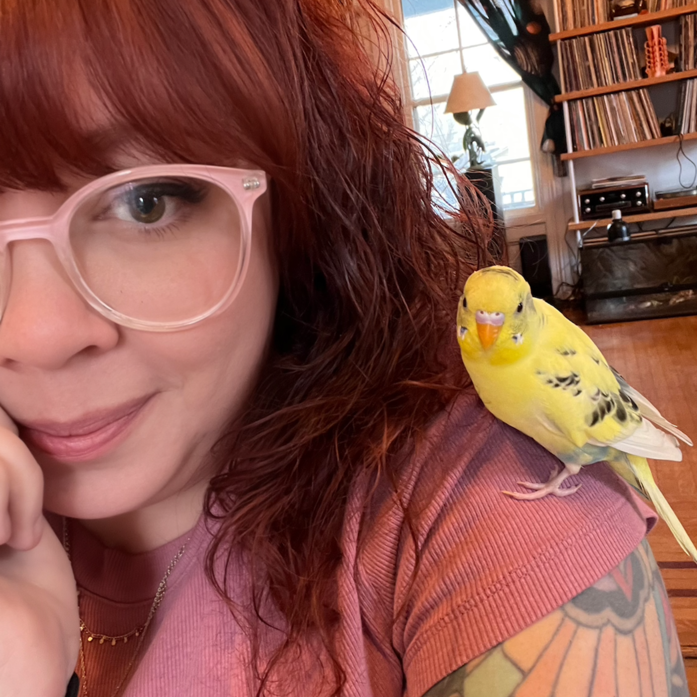 Woman with glasses and red hair has a yellow parakeet perched on her shoulder.