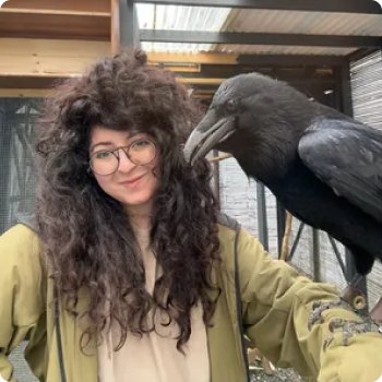 Woman with curly brown hair and glasses smiling next to a large black raven perched on a railing.