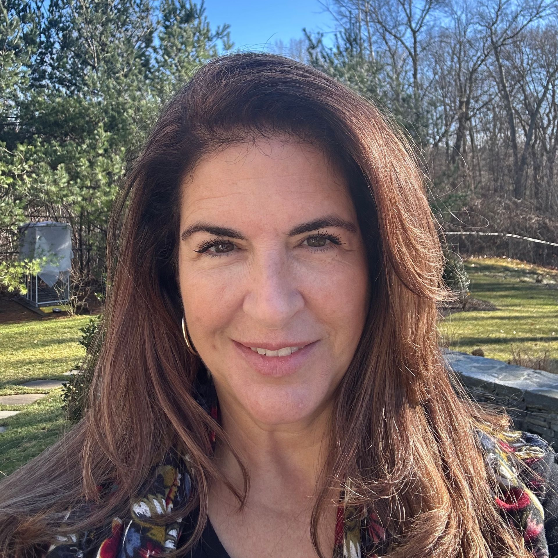 Woman with long brown hair smiles, outdoors. Trees and sunlight in background.