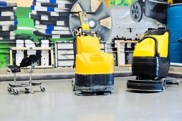 Yellow and black floor-cleaning machines in a workshop, with stacked tires and car wheels in the background