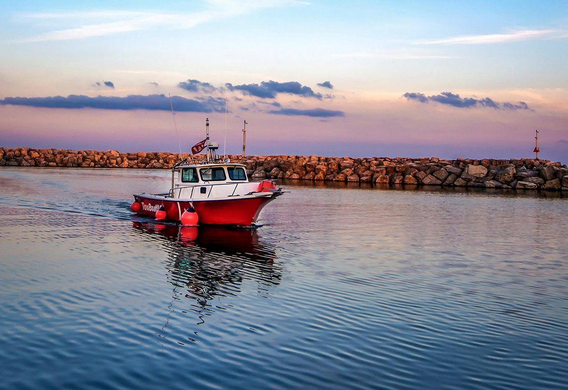 Red fishing boat floating on calm water near a rocky breakwater at sunset.