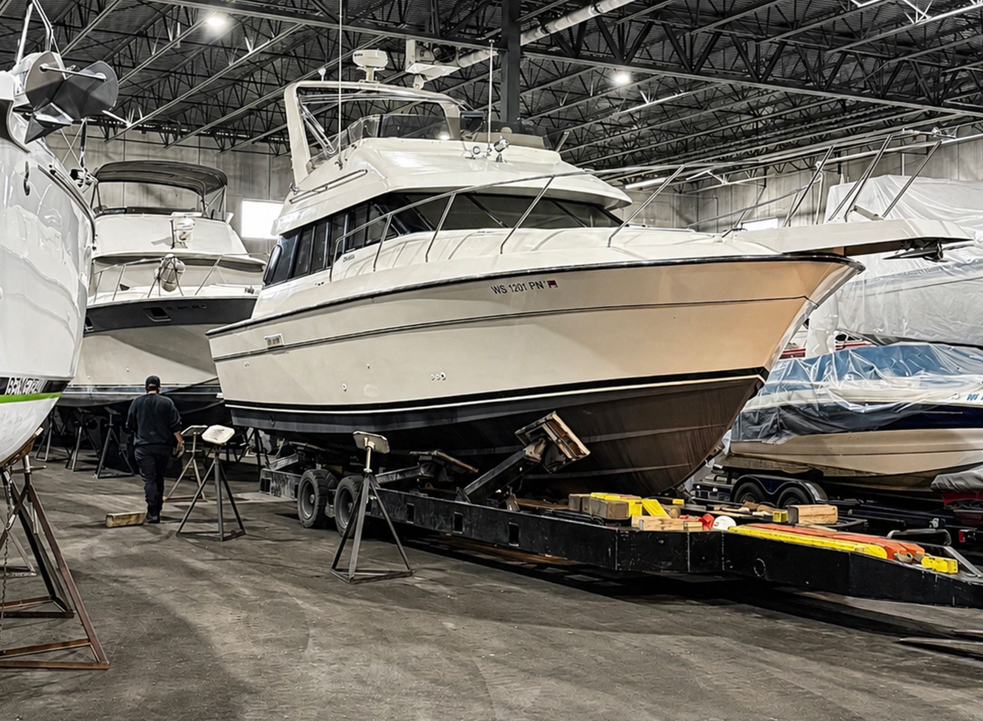 White motor yacht on stands inside a shipyard hangar, with workers and equipment nearby