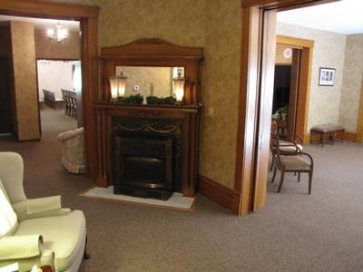 Fireplace in a room with doorway to another room; beige walls, carpet, chairs.