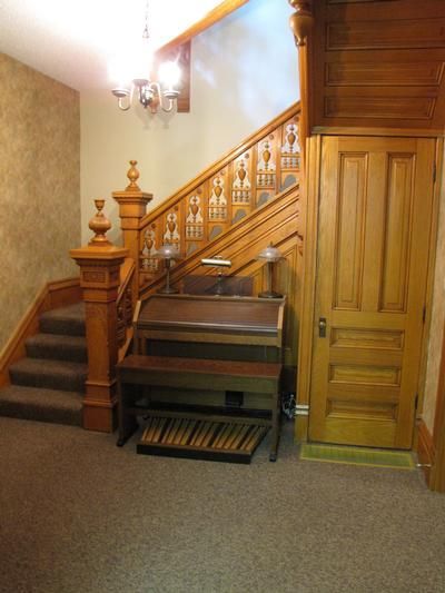 Wooden staircase with an organ in an entryway. The walls are wallpapered.