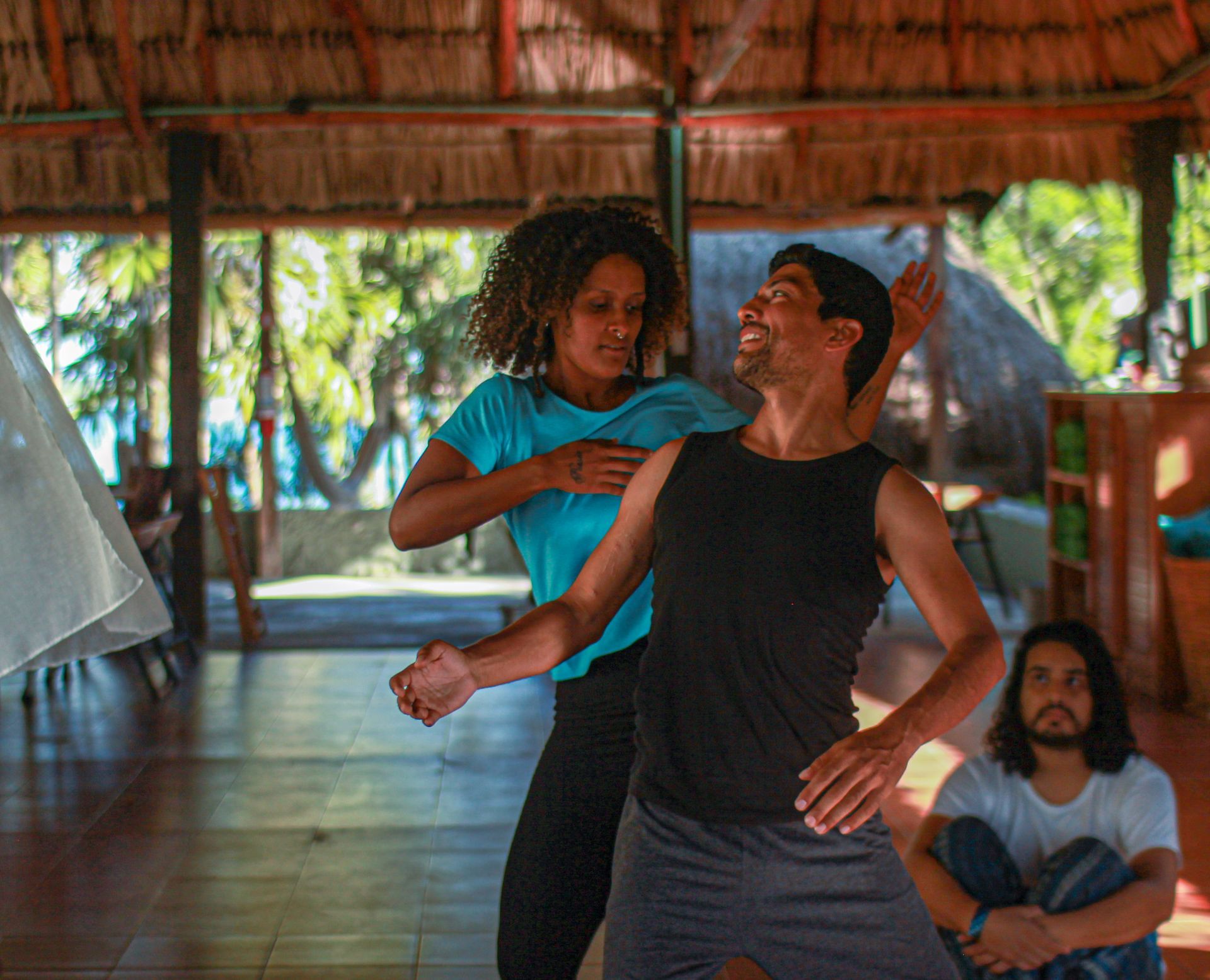 Dos personas bailan; una mujer con camiseta azul guía a un hombre con camiseta negra sin mangas. Un hombre observa. Estructura de madera, vista a la playa.