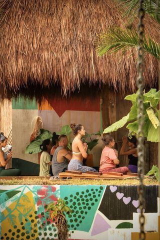Personas en clase de yoga bajo un techo de paja. Mural colorido, ambiente tropical. Manos en posición de oración.
